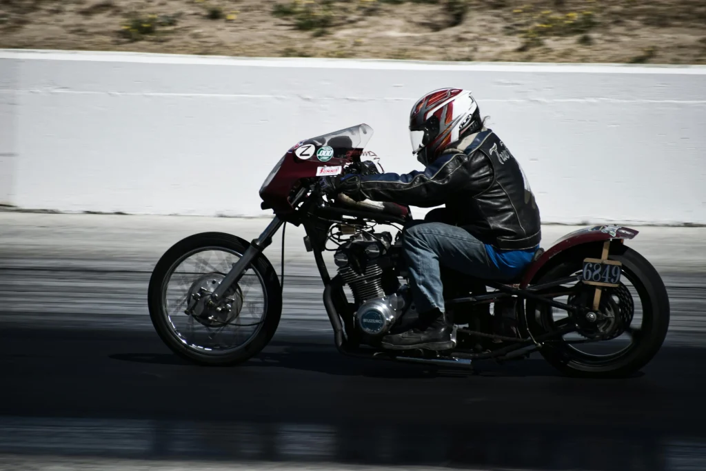 Motociclista compitiendo en pista con motocicleta estilo vintage dragster, usando casco integral y chaqueta negra.