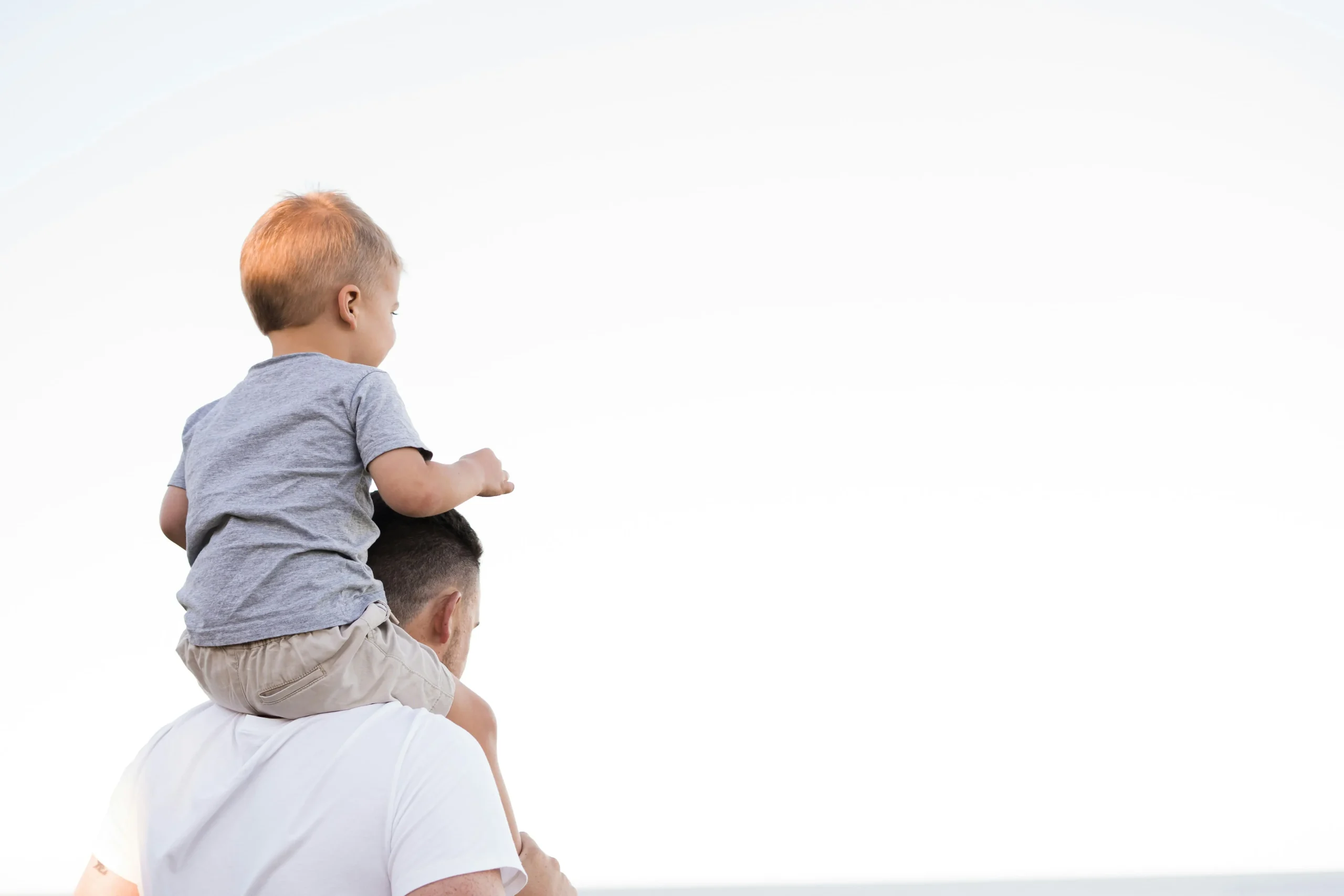 Niño pequeño feliz sobre los hombros de su padre al aire libre durante un día soleado.
